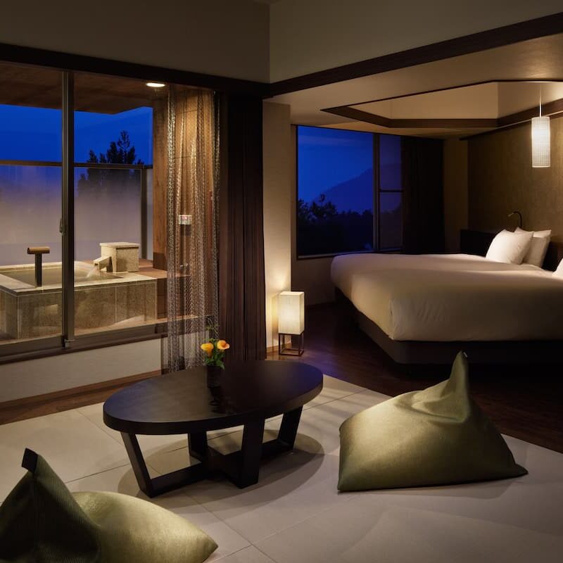 Detail suasana kamar tidur suite hotel premium di Jepang dengan desain modern dan sentuhan tradisional, menampilkan tempat tidur double yang nyaman dengan sprei putih, lampu tidur berdesain minimalis, area duduk dengan meja kopi oval dan bantal duduk lantai, serta kolam pemandian onsen pribadi dalam ruangan di dekat jendela besar yang menawarkan pemandangan pegunungan dan langit senja yang dramatis dalam wisata premium jepang 