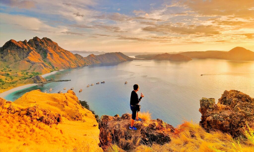 A hiker standing on a rocky viewpoint at Padar Island enjoying a spectacular sunrise over the iconic curved beaches in Komodo National Park.