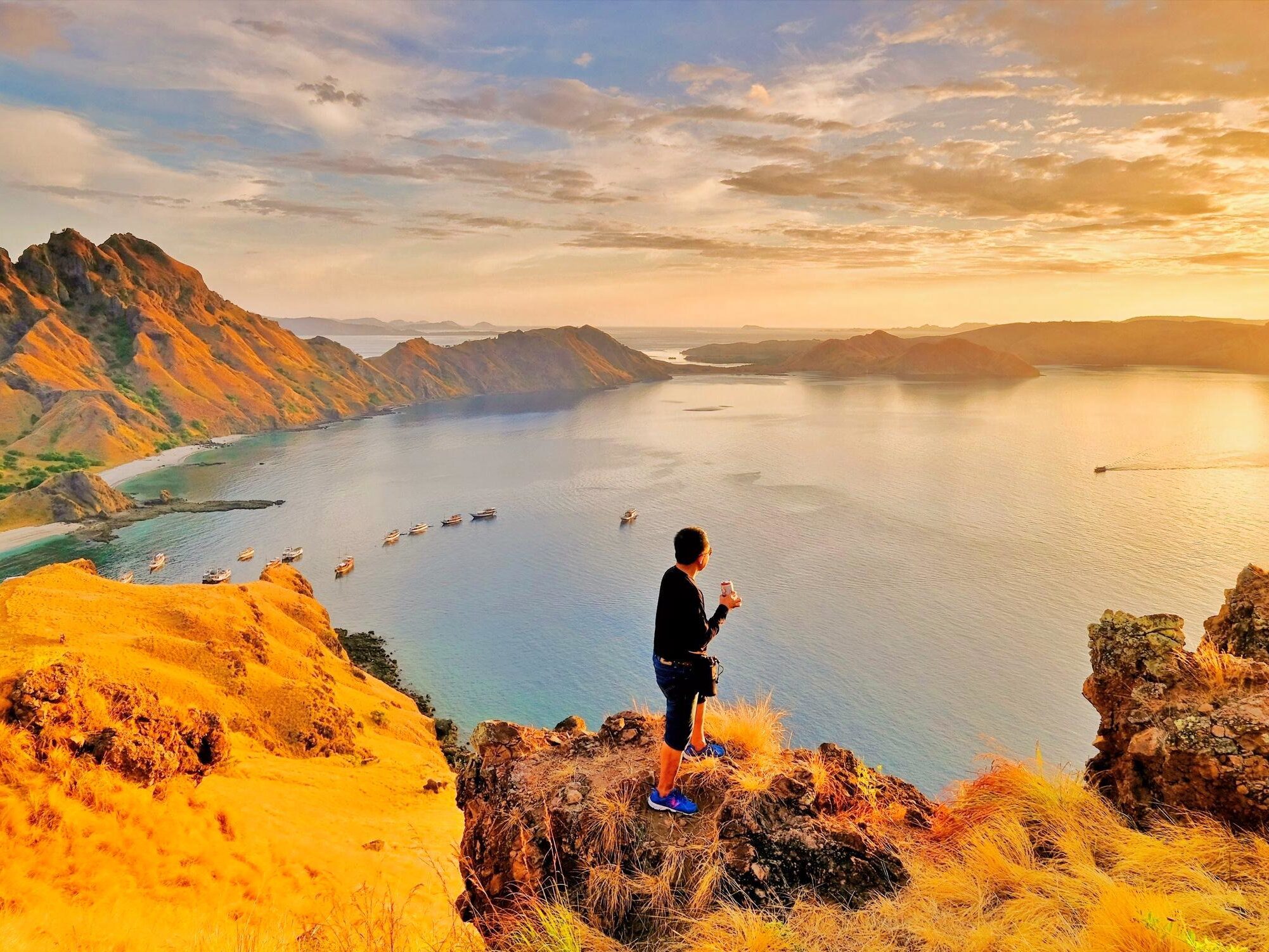 A hiker standing on a rocky viewpoint at Padar Island enjoying a spectacular sunrise over the iconic curved beaches in Komodo National Park.