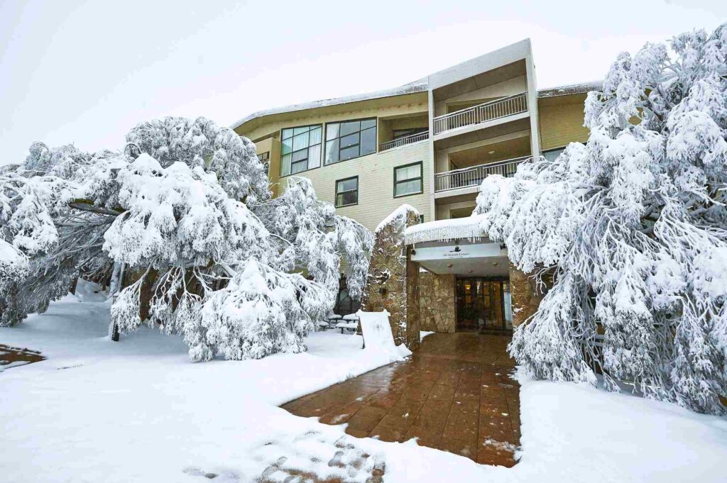 Pemandangan eksterior Mt Buller Chalet Hotel & Suites yang diselimuti salju tebal dan pohon-pohon beku, menawarkan pengalaman menginap musim dingin yang autentik dan eksklusif.