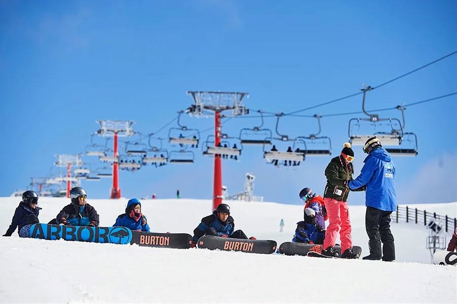 Peserta kursus snowboarding sedang beristirahat di lereng salju Mt. Buller yang cerah di bawah langit biru, lengkap dengan fasilitas kursi gantung ski di latar belakang.