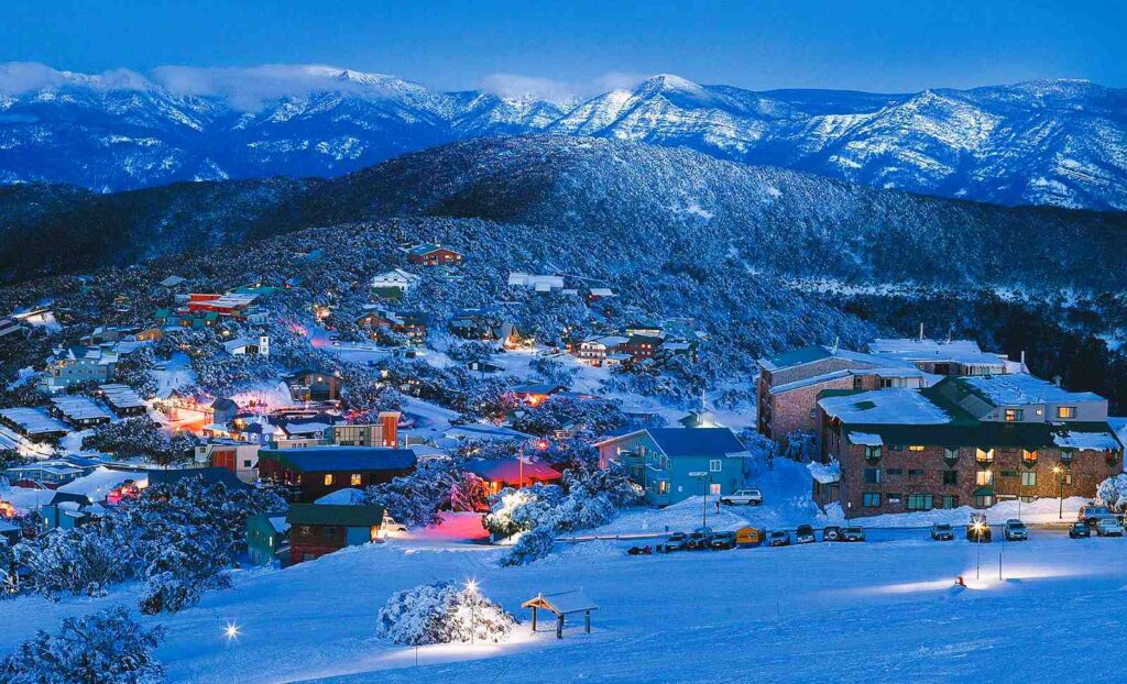 Pemandangan malam hari di Mt. Buller Alpine Village, Australia, menampilkan deretan penginapan yang tertutup salju dengan pendaran lampu desa yang hangat di tengah suasana musim dingin yang magis di private trip winter australia
