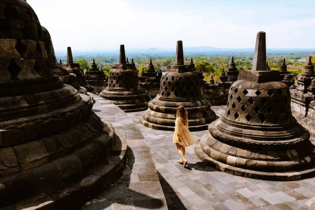 A woman in a yellow dress walks among the large, ancient stone bell-shaped stupas of Borobudur Temple under a clear sky, overlooking the lush green landscape of Central Java, Indonesia.