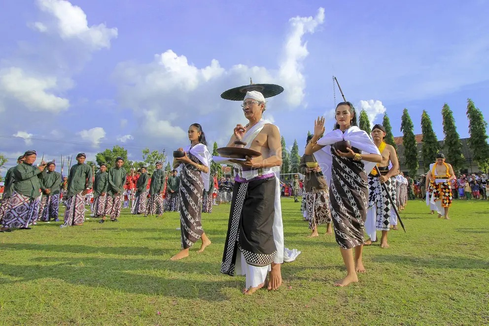 A documentary-style photograph of a traditional Javanese cultural procession on a grassy field. A central group, including a man with an intricate headdress and offering bowl, walks barefoot, flanked by men and women in traditional clothing, with spectators and pine trees in the background under a blue sky.