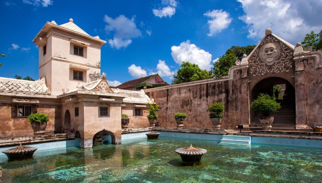 A historic Javanese water palace complex under a cloudy blue sky. A large, clear pool of water is surrounded by ancient stone architecture, a multi-story tower on the left, and an elaborate gateway with a carved monster face on the right leading up stairs.